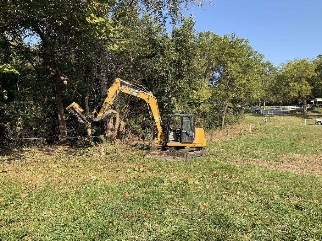 A Cat excavator clears underbrush and shrubs away from a fence line.